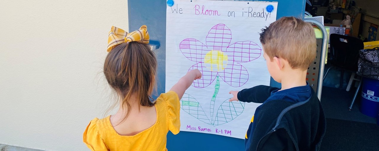 Two kindergarten students pointing to an iReady progress poster that has a partially coloerd in flower an says &#34;Blooming on i-Ready&#34;.